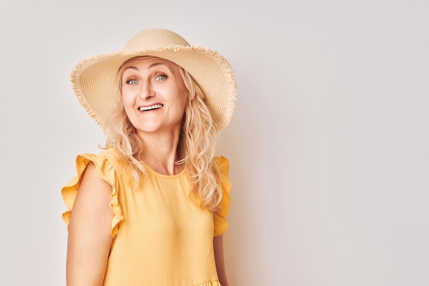 Ritratto di felice bella donna matura bionda in abito estivo giallo e cappello di paglia sorridente con gioia isolato su sfondo bianco studio Concetto di vacanze e viaggi
