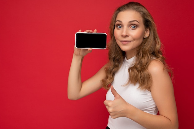 Ritratto di felice bella giovane donna bionda che indossa t-shirt bianca isolata su sfondo rosso con spazio copia che tiene smartphone che mostra telefono in mano con schermo vuoto per mockup guardando la fotocamera