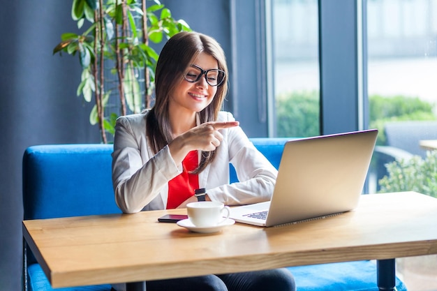 Ritratto di felice bella giovane donna bruna alla moda con gli occhiali seduto a guardare il suo schermo del computer portatile su puntamento videochiamata e sorriso a trentadue denti indoor studio girato sfondo caffè ufficio