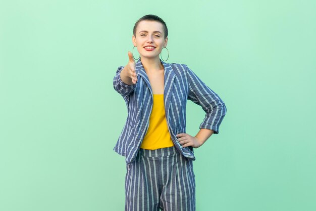 Ritratto di felice bello capelli corti giovane donna alla moda in abito a righe casual in piedi con un sorriso a trentadue denti e dando la mano al saluto. studio indoor girato isolato su sfondo verde chiaro