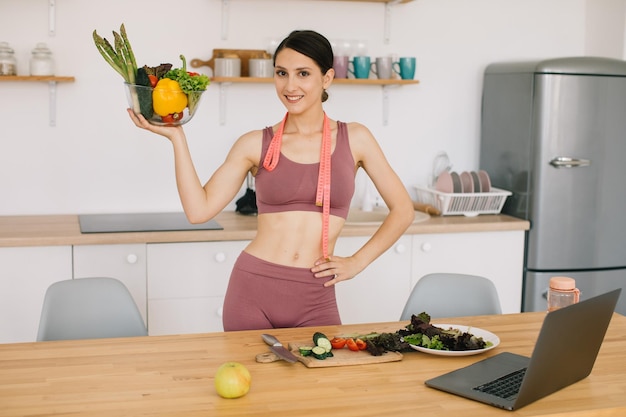 Ritratto di felice blogger donna sportiva che tiene piatto di verdure fresche e conduce una videoconferenza su un'alimentazione sana sul computer portatile in cucina