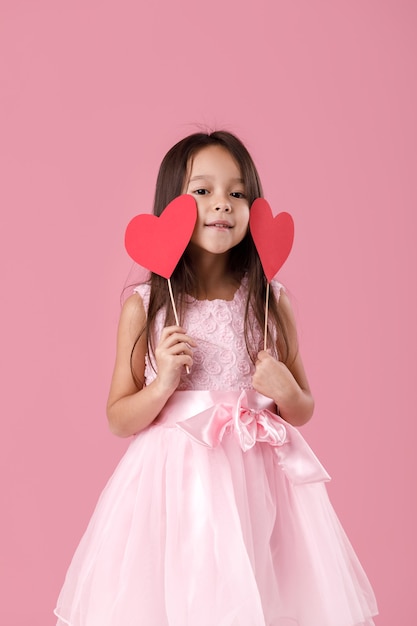 Ritratto di felice carino piccolo bambino ragazza in un vestito rosa che tiene due cuori di carta su uno sfondo rosa. San Valentino