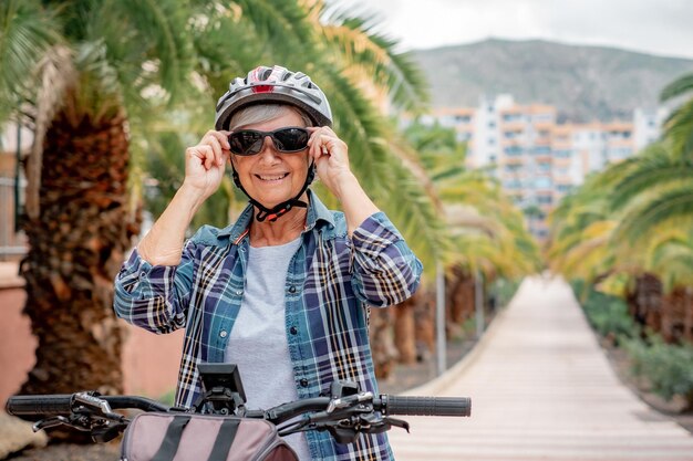 Ritratto di felice ciclista senior con casco e occhiali da sole nel parco pubblico che corre con la sua bicicletta elettrica Stile di vita sano per i pensionati e mobilità sostenibile