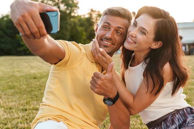 Ritratto di felice coppia adulta uomo e donna che mostra il pollice in su e scatta una foto selfie sul cellulare mentre è seduto sull'erba nel parco