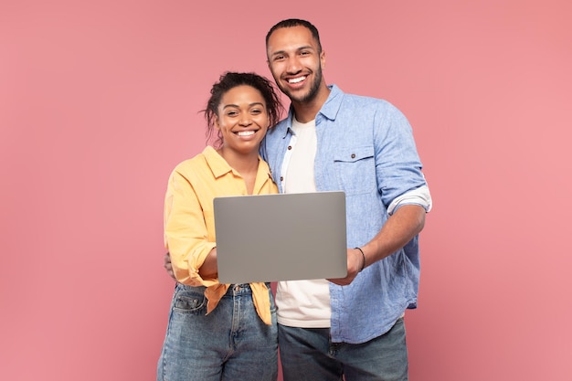 Ritratto di felice coppia afroamericana che tiene e utilizza il laptop in posa su sfondo rosa e sorride alla telecamera