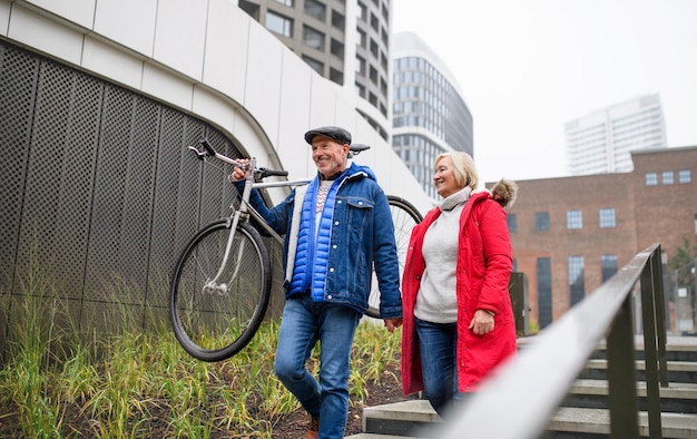 Ritratto di felice coppia senior con bicicletta a piedi in città, tenendosi per mano.