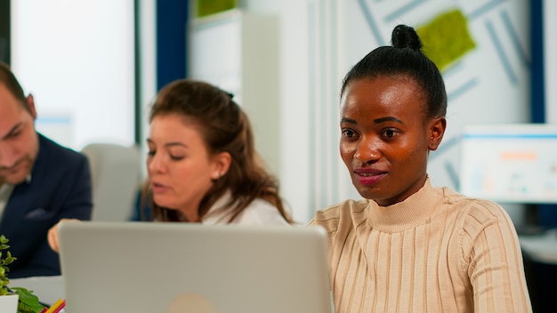 Ritratto di felice donna d'affari africana che legge buone notizie sul computer portatile seduto alla scrivania in un ufficio di avvio occupato mentre un team diversificato analizza i dati statistici. Team multietnico al lavoro su un nuovo progetto