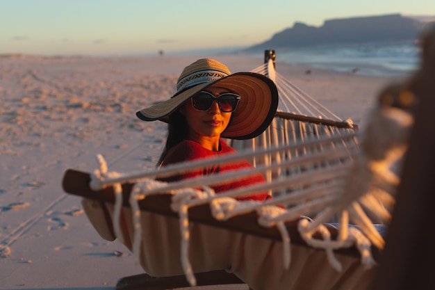 Ritratto di felice donna di razza mista in vacanza al mare che guarda alla telecamera seduta in amaca al tramonto. sano tempo libero all'aria aperta in riva al mare.