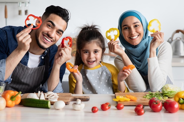 Ritratto di felice famiglia araba con la piccola figlia che tiene le fette di peperone mentre posano in cucina insieme, allegra famiglia mediorientale di tre che si divertono mentre cucinano cibo sano a casa