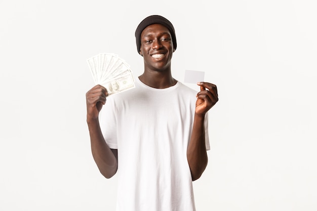 Ritratto di felice, fortunato maschio afro-americano in berretto, sorridente allegro mentre mostra la carta di credito con i soldi
