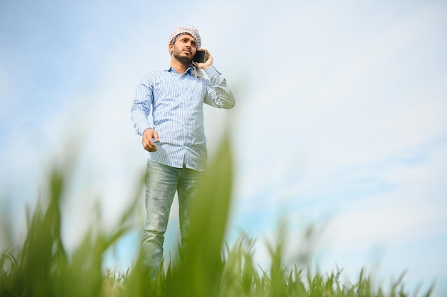 Ritratto di felice giovane agricoltore indiano in piedi in terreno agricolo circondato da raccolto che tiene smartphone che parla con qualcuno sorridente maschio del villaggio con il telefono cellulare nel concetto di tecnologia sul campo