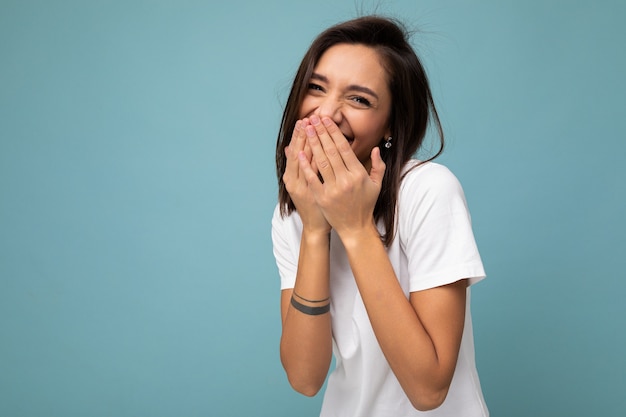 Ritratto di felice giovane bella signora brunet positiva con emozioni sincere che indossa t-shirt bianca casual per mockup isolato su sfondo blu con spazio copia e ridendo che copre la bocca