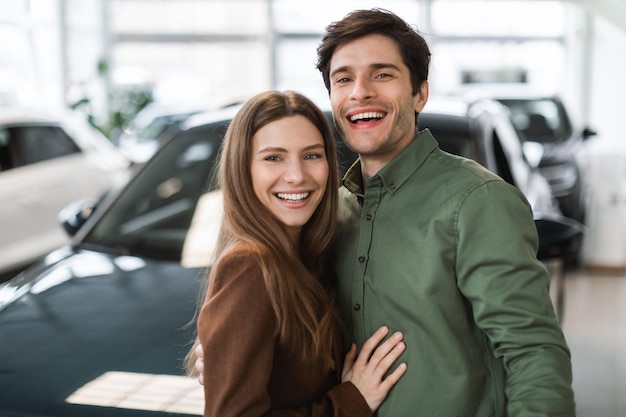 Ritratto di felice giovane coppia caucasica in posa e sorridente alla telecamera vicino alla nuova auto presso la concessionaria