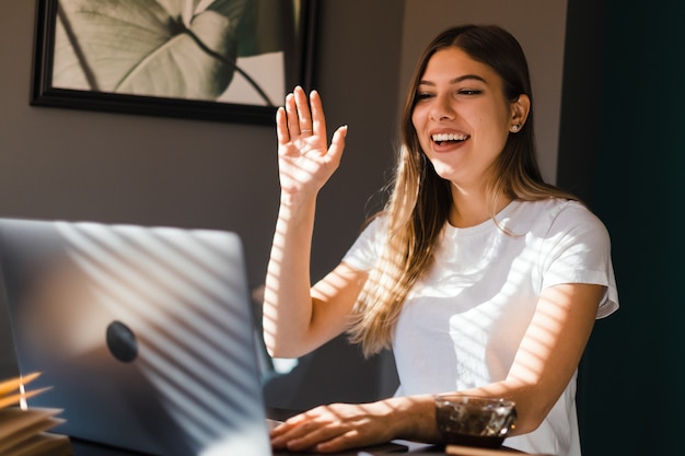 Ritratto di felice giovane donna bruna seduta al tavolo, guardando lo schermo del laptop in videochiamata e saluto con agitando