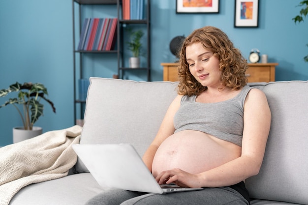 Ritratto di felice giovane donna incinta seduta sul divano e guardando il computer portatile