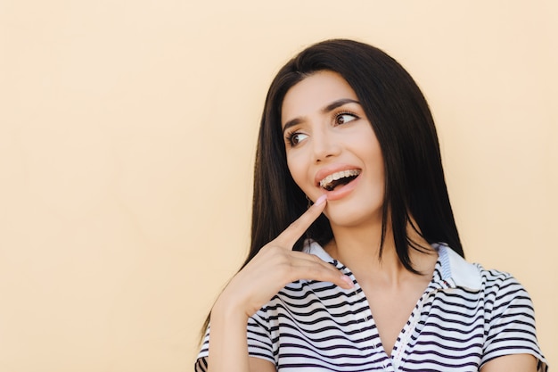 Ritratto di felice giovane femmina con lunghi capelli lisci scuri, ha un ampio sorriso, indica alla sua bocca