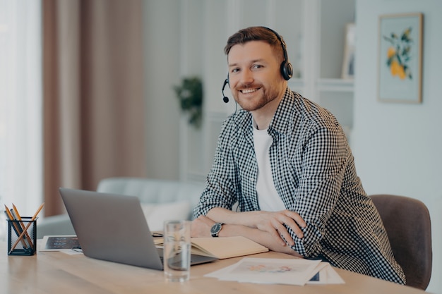 Ritratto di felice giovane libero professionista maschio seduto nel suo moderno e accogliente posto di lavoro e lavorando al computer portatile, ragazzo allegro che sorride alla macchina fotografica e si gode il lavoro a distanza. Concetto di freelance