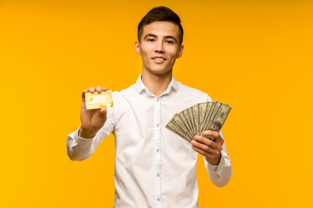 Ritratto di felice giovane uomo asiatico tenendo la carta di credito e denaro in mano sorridendo e guardando la telecamera su sfondo giallo isolato sensazione positiva e godere - immagine