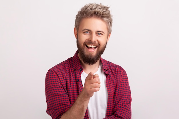 Ritratto di felice maschio barbuto in camicia rossa che sorride sinceramente alla telecamera e fa cenno con il dito. Studio indoor girato isolato su sfondo grigio