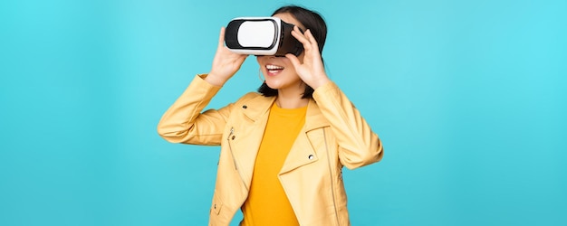 Ritratto di felice modello femminile asiatico utilizzando auricolare VR sorridente e ridendo in occhiali per realtà virtuale in piedi su sfondo blu