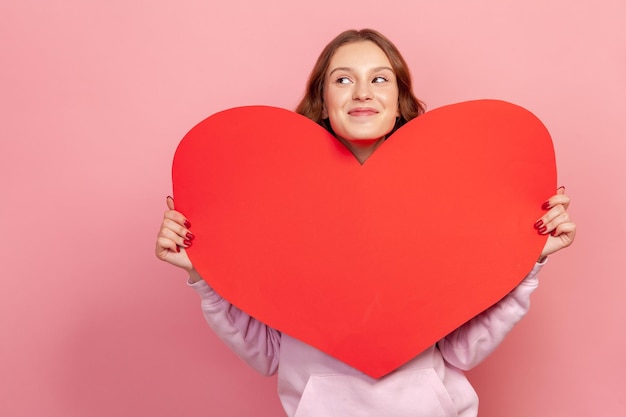 Ritratto di felice ragazza adolescente in felpa con cappuccio che tiene un enorme cuore di carta e guardando al lato innamorarsi del simbolo del giorno di San Valentino Colpo in studio al coperto isolato su sfondo rosa