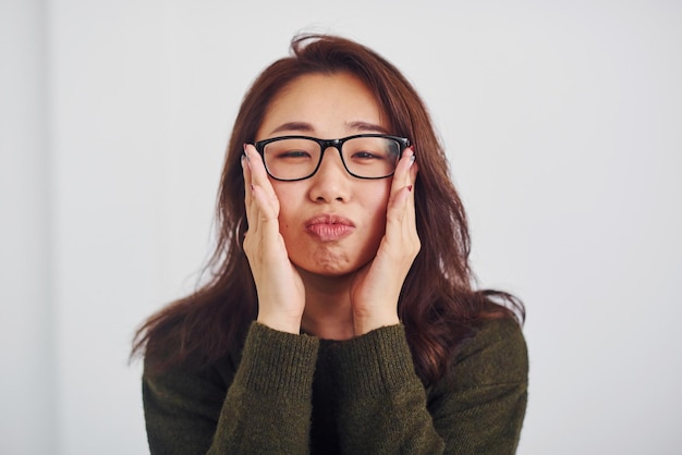 Ritratto di felice ragazza asiatica in occhiali che in piedi al chiuso in studio su sfondo bianco.