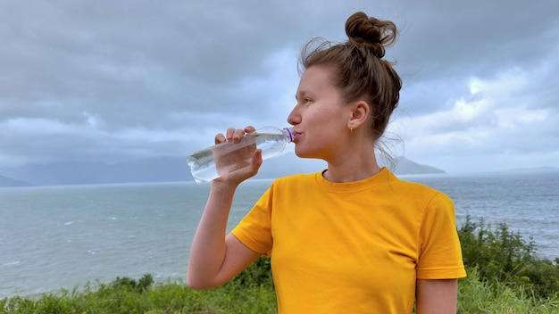 Ritratto di felice ragazza assetata positiva giovane bella donna sta bevendo pura acqua minerale fresca dalla bottiglia di plastica sullo sfondo naturale dell'oceano di mare nel paese tropicale tenendo la bottiglia in mano