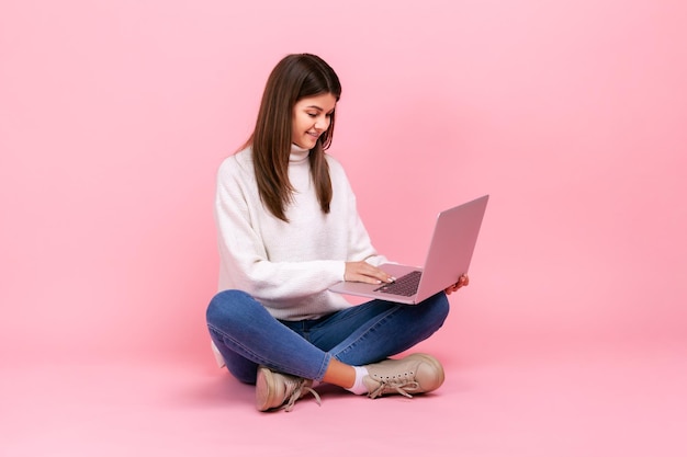 Ritratto di felice ragazza bruna seduta sul pavimento con le gambe incrociate e lavorando su laptop freelancer che indossa un maglione bianco in stile casual Studio indoor girato isolato su sfondo rosa