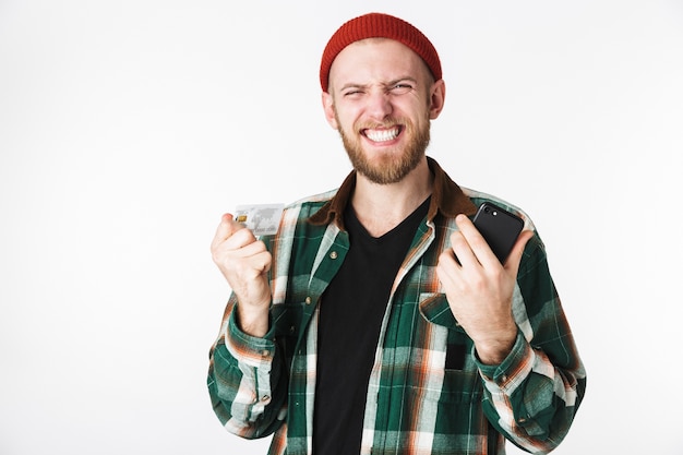 Ritratto di felice ragazzo barbuto che indossa un cappello e camicia a quadri sorridente, mentre in piedi isolato su sfondo bianco