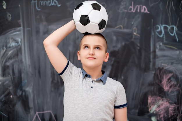 ritratto di felice ragazzo carino che si diverte a tenere un pallone da calcio in testa mentre si trova davanti alla lavagna nera