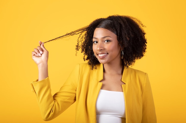 Ritratto di felice signora afroamericana che tocca i capelli e flirta guardando la fotocamera e sorridendo