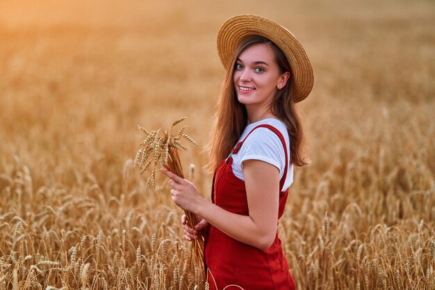 Ritratto di felice sorridente attraente carina libera giovane femmina che indossa cappello di paglia e denim in piedi nel campo di grano giallo dorato e godersi la bella vita momento di libertà in estate