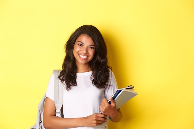 Ritratto di felice studentessa universitaria afro-americana, con in mano taccuini e zaino, sorridente e in piedi su sfondo giallo.