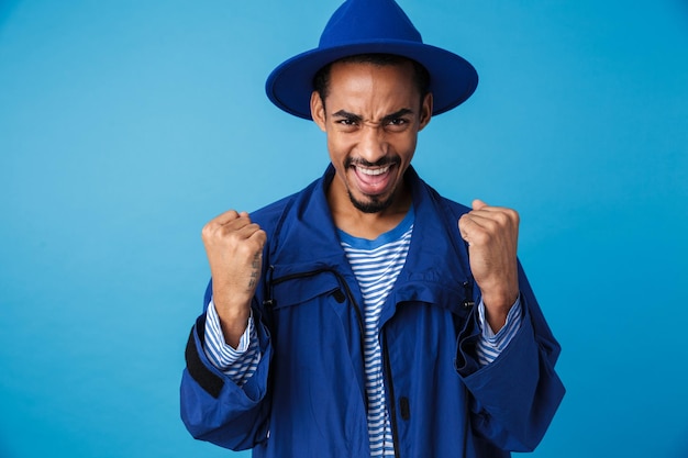 Ritratto di felice uomo afroamericano che indossa un cappello che celebra il successo e sorride isolato su blue