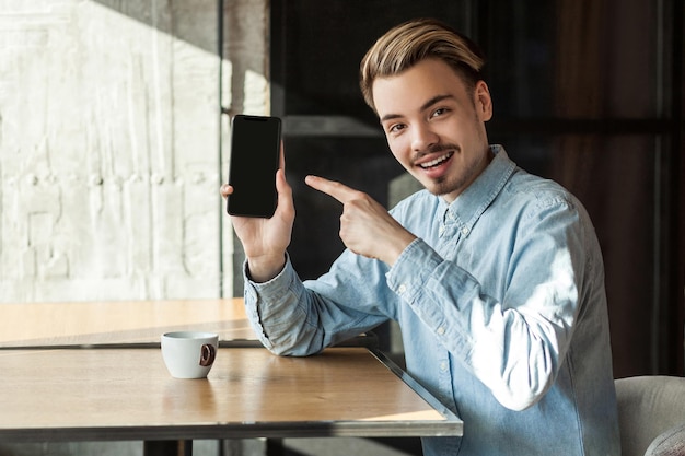 Ritratto di felice uomo barbuto positivo in camicia blu denim seduto al bar, tenendo il telefono e puntando il dito sul display mobile con un sorriso a trentadue denti e una faccia soddisfatta. Al chiuso, stile di vita