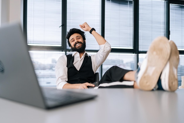 Ritratto di felice uomo d'affari indiano seduto alla scrivania dell'ufficio con i piedi sul tavolo con il computer portatile lavoro finito lavoro completato tutte le attività rilassanti dopo una dura giornata lavorativa sorridente guardando la fotocamera