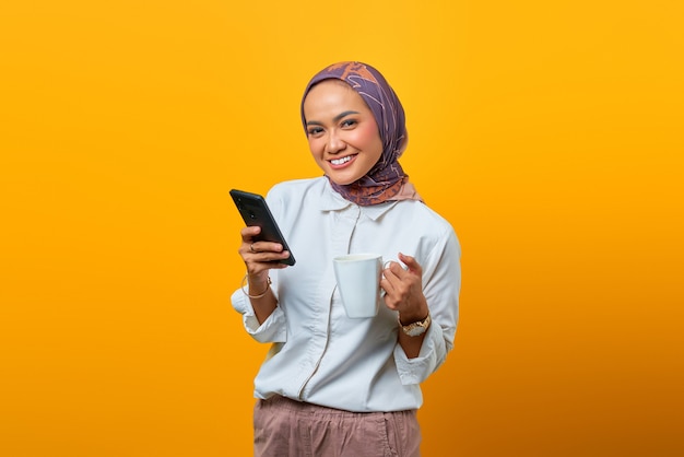 Ritratto di felicità Donna asiatica che si rilassa tenendo la tazza e usa il telefono cellulare su sfondo giallo