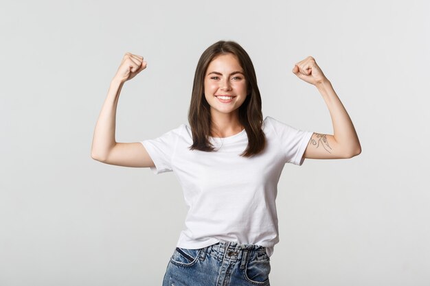 Ritratto di fiduciosa ragazza attraente flessione bicipite per mostrare i punti di forza, sorridente impertinente su bianco.