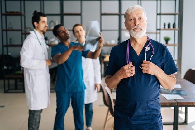 Ritratto di fiducioso chirurgo anziano in uniforme blu in piedi nell'ufficio della riunione medica guardando la fotocamera