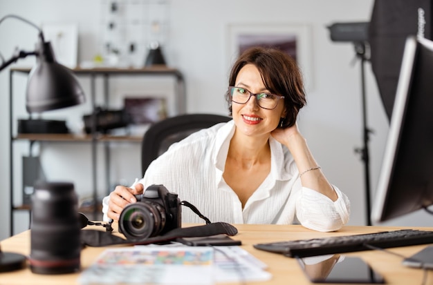 Ritratto di fotografa sorridente in abbigliamento elegante seduto nel proprio ufficio con fotocamera e computer moderni Donna creativa sul posto di lavoro