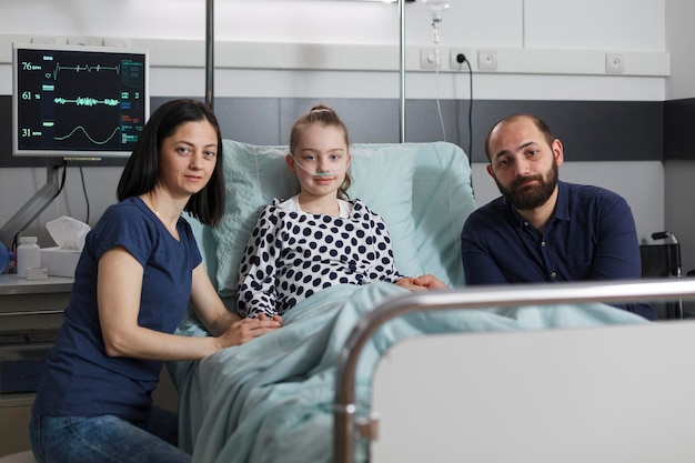 Ritratto di genitori premurosi seduti accanto alla figlia malata ricoverata che riposa nel letto del paziente della struttura sanitaria dei bambini mentre guarda la fotocamera. Famiglia caucasica all'interno del reparto pediatrico dell'ospedale.