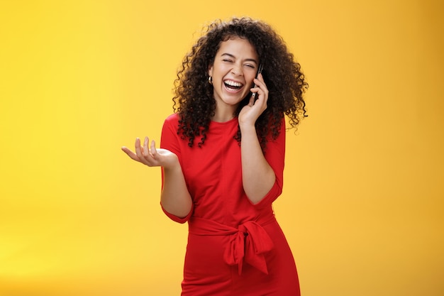 Ritratto di gioiosa affascinante femmina europea in abito rosso con i capelli ricci che ride ad alta voce mentre spettegola con un amico tramite smartphone chiudi gli occhi mentre ridacchia gesticolando, ascoltando uno scherzo tramite il telefono cellulare.