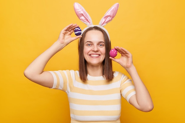 Ritratto di gioiosa donna ottimista positiva che indossa orecchie da coniglio tenendo le uova di pasqua isolate su sfondo giallo guardando la fotocamera con un sorriso che celebra la vacanza