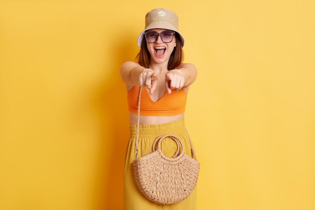 Ritratto di gioiosa giovane donna sorridente che indossa abiti estivi con borsa di paglia in piedi isolato su sfondo giallo che punta alla fotocamera facendo scelta dicendo bisogno di te