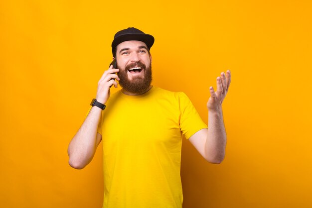 Ritratto di gioioso hipster barbuto uomo in piedi sopra il muro giallo e parlando sullo smartphone