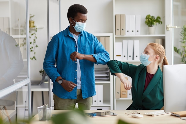 Ritratto di giovane afro-americano che indossa la maschera urtando i gomiti con il collega di sesso femminile come saluto contactless in post pandemia office