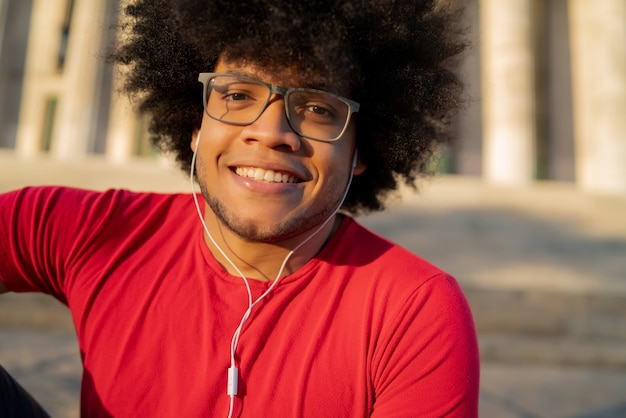 Ritratto di giovane afro latino uomo ascoltando musica con gli auricolari e rilassarsi mentre è seduto sulle scale all'aperto. Concetto urbano.