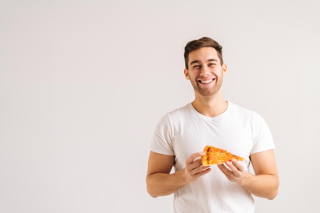 Ritratto di giovane allegro che tiene in mano una fetta di deliziosa pizza, in piedi in posa su sfondo bianco isolato. Studio girato di bello studente maschio felice che mangia un pasto gustoso.