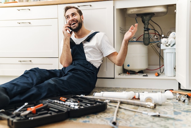 Ritratto di giovane allegro uomo sorridente idraulico lavoro in uniforme al chiuso parlando dal telefono cellulare.