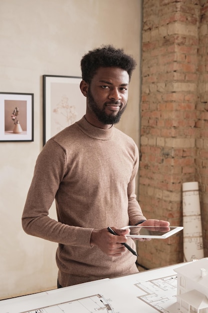 Ritratto di giovane architetto nero sorridente con la barba in piedi al tavolo con progetto e utilizzando tablet mentre elabora il design dell'edificio